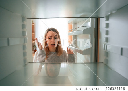 Disappointed about an empty fridge with no food in it. Beautiful young woman is on the kitchen 126323118