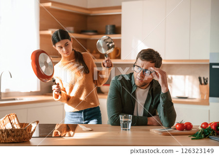 Woman is holding the pan and wants to hit a guy. Young couple is on the kitchen together Woman is holding the pan and wants to hit a guy. Young couple is on the kitchen together 126323149