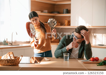 Woman is holding the pan and wants to hit a guy. Young couple is on the kitchen together Woman is holding the pan and wants to hit a guy. Young couple is on the kitchen together 126323150