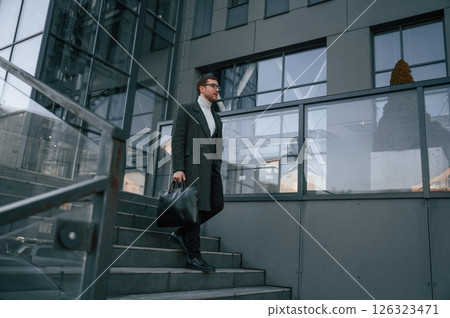 Holding the bag. Handsome man in formal clothes is outdoor near the business building 126323471