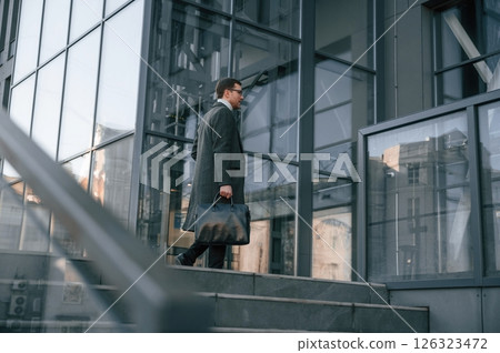 Holding the bag. Handsome man in formal clothes is outdoor near the business building 126323472
