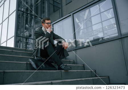 Sitting on the stairs and smoking. Handsome man in formal clothes is outdoor near the business building 126323473