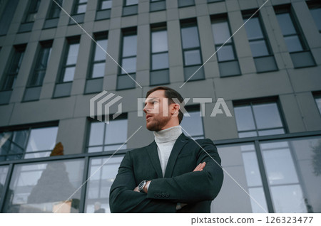 Standing with arms crossed. Handsome man in formal clothes is outdoor near the business building 126323477