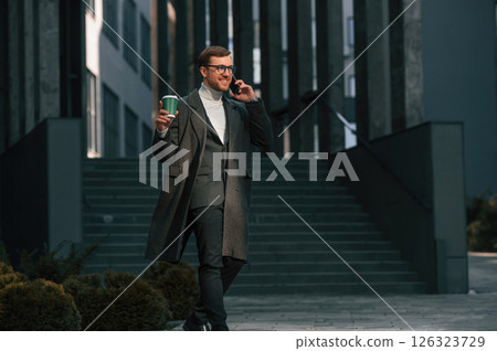 Amazing sunlight, holding smartphone and cup of drink. Handsome man in formal clothes is outdoor near the business building 126323729