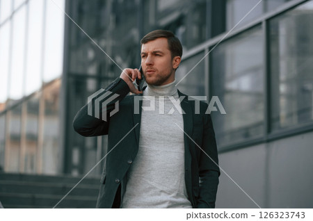 Having conversation by phone. Handsome man in formal clothes is outdoor near the business building 126323743