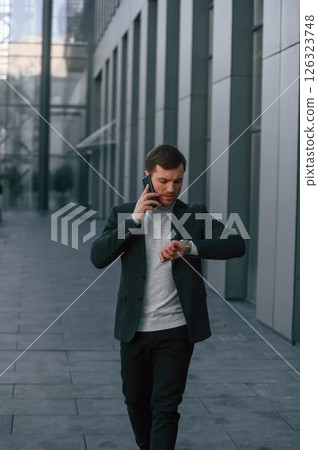 Having conversation by phone. Handsome man in formal clothes is outdoor near the business building 126323748