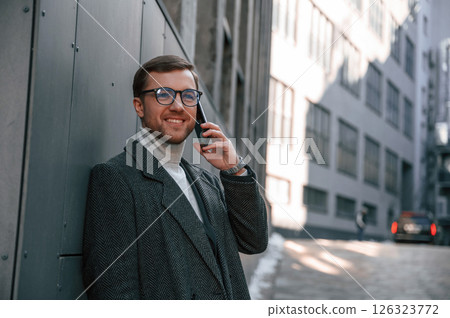 Black colored smartphone in hands. Handsome man in formal clothes is outdoor near the business building Black colored smartphone in hands. Handsome man in formal clothes is outdoor near the business building 126323772