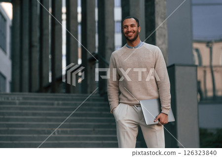 Positive handsome black man is outdoors near the business building holding laptop 126323840