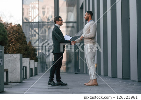 Doing handshake, greetings gesture. Businessman in formal clothes is with black man outdoors in the city 126323961