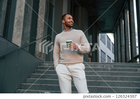 With cup of drink. Handsome black man is outdoors near the business building 126324307