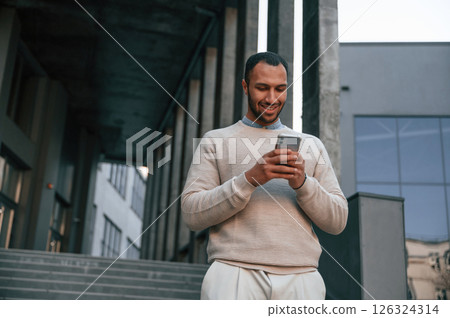 Enjoying social media by using smartphone. Handsome black man is outdoors near the business building 126324314
