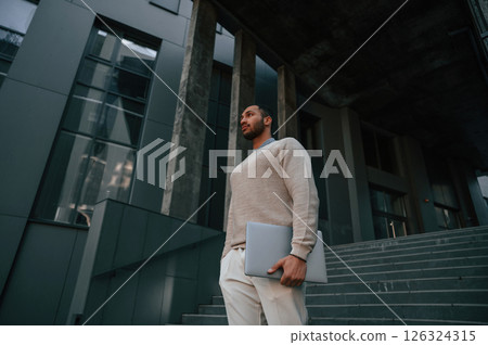 Positive handsome black man is outdoors near the business building holding laptop 126324315