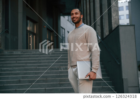 Positive handsome black man is outdoors near the business building holding laptop Positive handsome black man is outdoors near the business building holding laptop 126324318