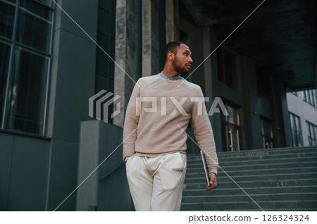 Front view. Standing with laptop in hands. Handsome black man is outdoors near the business building Front view. Standing with laptop in hands. Handsome black man is outdoors near the business building 126324324