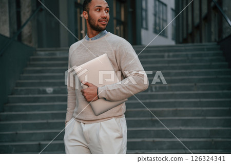 Holding laptop in hand. Handsome black man is outdoors near the business building 126324341