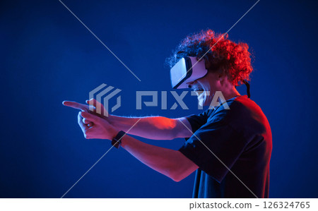 Pistol gesture by hands. In VR glasses. Young man with curly hair is indoors illuminated by neon lighting 126324765