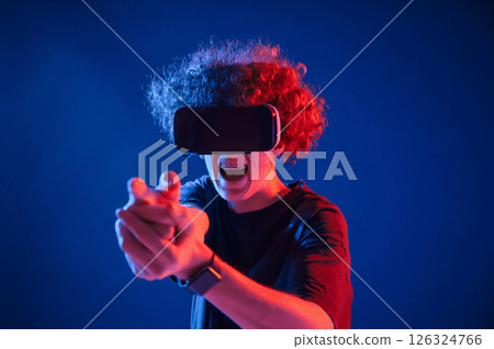 Pistol gesture by hands. In VR glasses. Young man with curly hair is indoors illuminated by neon lighting 126324766