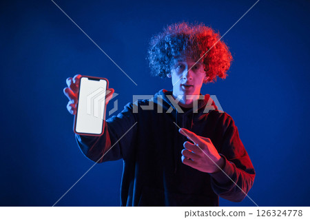 Holding smartphone and showing it to the camera. Young man with curly hair is indoors illuminated by neon lighting Holding smartphone and showing it to the camera. Young man with curly hair is indoors illuminated by neon lighting 126324778