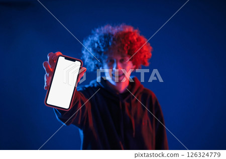 Holding smartphone and showing it to the camera. Young man with curly hair is indoors illuminated by neon lighting Holding smartphone and showing it to the camera. Young man with curly hair is indoors illuminated by neon lighting 126324779