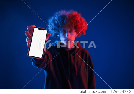 Holding smartphone and showing it to the camera. Young man with curly hair is indoors illuminated by neon lighting Holding smartphone and showing it to the camera. Young man with curly hair is indoors illuminated by neon lighting 126324780