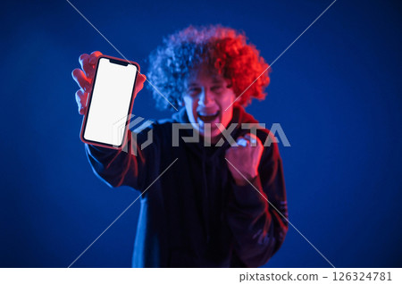 Showing info on the smartphone. Young man with curly hair is indoors illuminated by neon lighting 126324781