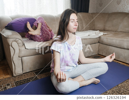 Young mother tries to meditate and do a home workout while her playful daughter gets in her way. Interrupted yoga at home and parenting reality. Mom and daughter having fun Young mother tries to meditate and do a home workout while her playful daughter gets in her way. Interrupted yoga at home and parenting reality. Mom and daughter having fun 126326194