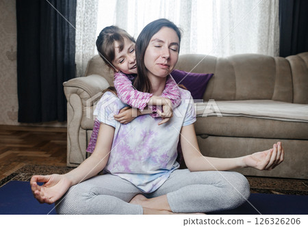 Young mother tries to meditate and do a home workout while her playful daughter gets in her way. Interrupted yoga at home and parenting reality. Mom and daughter having fun Young mother tries to meditate and do a home workout while her playful daughter gets in her way. Interrupted yoga at home and parenting reality. Mom and daughter having fun 126326206
