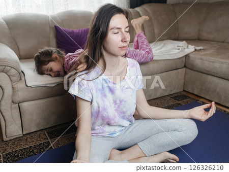 Young mother tries to meditate and do a home workout while her playful daughter gets in her way. Interrupted yoga at home and parenting reality. Mom and daughter having fun Young mother tries to meditate and do a home workout while her playful daughter gets in her way. Interrupted yoga at home and parenting reality. Mom and daughter having fun 126326210