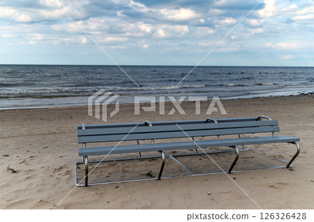 A solitary bench on a sandy beach with calm waves under a cloudy sky in the late afternoon 126326428