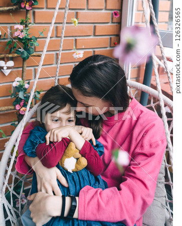 Mother and daughter hugging in a wicker garden swing surrounded by flowers. The concept of a happy childhood and loving caring parents 126326565