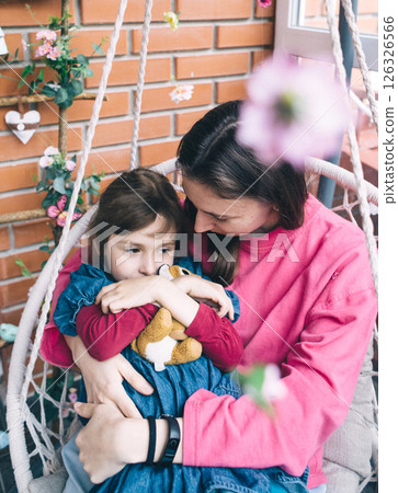 Mother and daughter hugging in a wicker garden swing surrounded by flowers. The concept of a happy childhood and loving caring parents 126326566