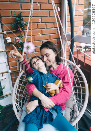 Mother and daughter hugging in a wicker garden swing surrounded by flowers. The concept of a happy childhood and loving caring parents 126326567