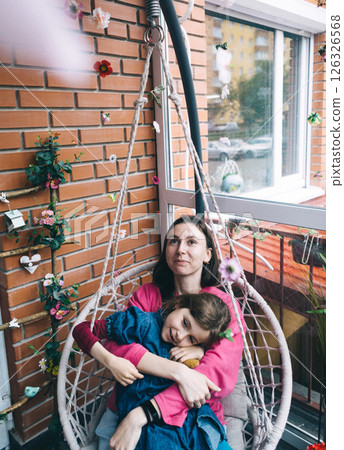 Mother and daughter hugging in a wicker garden swing surrounded by flowers. The concept of a happy childhood and loving caring parents 126326568