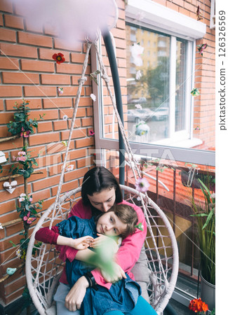 Mother and daughter hugging in a wicker garden swing surrounded by flowers. The concept of a happy childhood and loving caring parents 126326569
