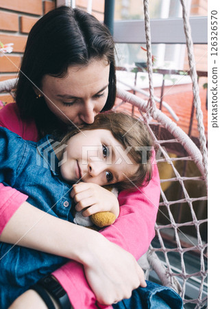 Mother and daughter hugging in a wicker garden swing surrounded by flowers. The concept of a happy childhood and loving caring parents 126326570