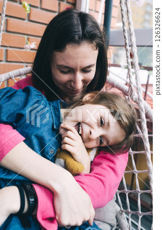 Mother and daughter hugging in a wicker garden swing surrounded by flowers. The concept of a happy childhood and loving caring parents 126326624