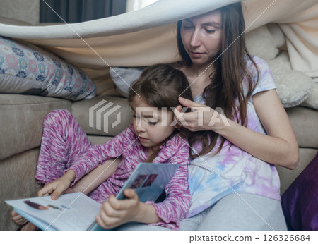Mother and daughter sit in a hut made of a blanket and read a book. Pleasant time with children, quiet time before bed. Mother and daughter sit in a hut made of a blanket and read a book. Pleasant time with children, quiet time before bed. 126326684