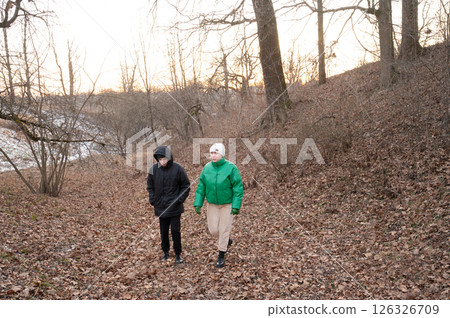 Two friends enjoy a peaceful winter walk along a leaf covered trail beneath bare trees during sunset in a serene countryside setting 126326709