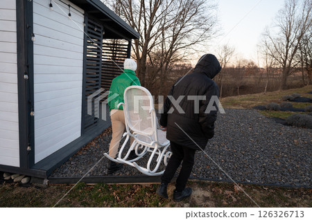 Two individuals transport a white rocking chair towards a cozy shed in the fading light of twilight, illustrating the quiet charm of early evening at a rural retreat 126326713
