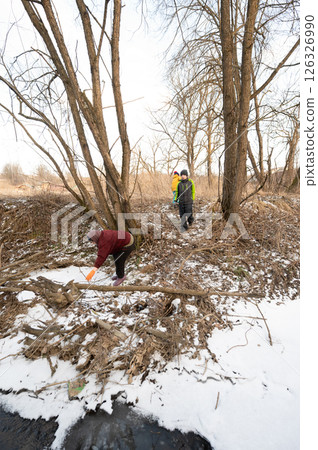 Exploring a snowy landscape by the river, individuals engaged in cleanup activities amidst winter foliage and bare trees under a clear sky 126326990