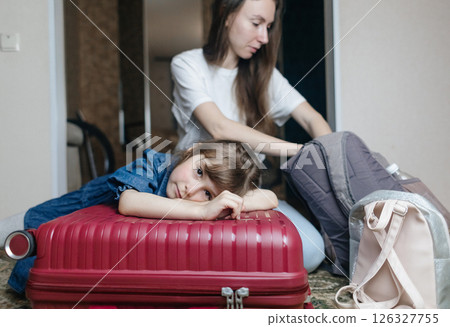 Tired 6 year girl lying on travel suitcase while mother is getting ready for trip. Cute child lost strength after packing bag and preparing for trip. Loving family, child care, home schooling concept 126327755