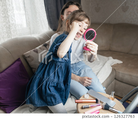 Daughter puts makeup on her mother face and plays around. Caring parent allows her child to paint her face. Happy childhood. Concept of loving family and fun. little girl doing her makeup 126327763