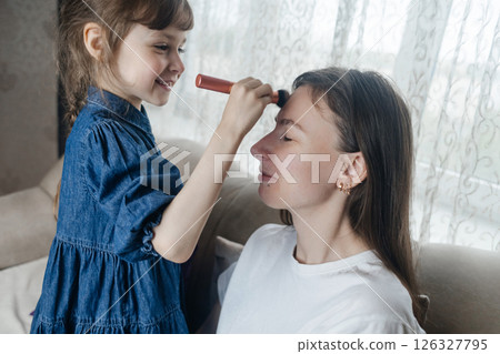 Daughter puts makeup on her mother face and plays around. Caring parent allows her child to paint her face. Happy childhood. Concept of loving family and fun. little girl doing her makeup Daughter puts makeup on her mother face and plays around. Caring parent allows her child to paint her face. Happy childhood. Concept of loving family and fun. little girl doing her makeup 126327795