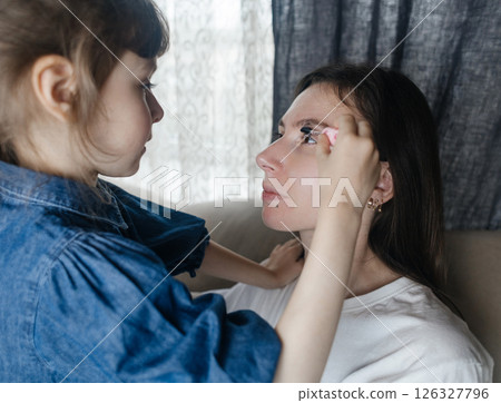 Daughter puts makeup on her mother face and plays around. Caring parent allows her child to paint her face. Happy childhood. Concept of loving family and fun. little girl doing her makeup 126327796