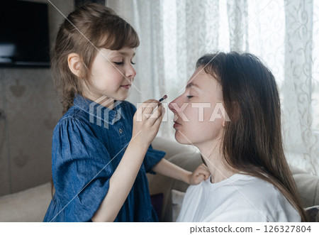 Daughter puts makeup on her mother face and plays around. Caring parent allows her child to paint her face. Happy childhood. Concept of loving family and fun. little girl doing her makeup Daughter puts makeup on her mother face and plays around. Caring parent allows her child to paint her face. Happy childhood. Concept of loving family and fun. little girl doing her makeup 126327804