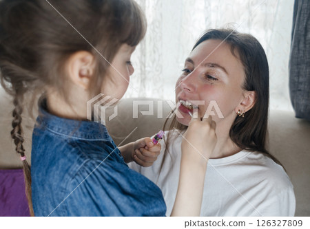 Daughter puts makeup on her mother face and plays around. Caring parent allows her child to paint her face. Happy childhood. Concept of loving family and fun. little girl doing her makeup Daughter puts makeup on her mother face and plays around. Caring parent allows her child to paint her face. Happy childhood. Concept of loving family and fun. little girl doing her makeup 126327809