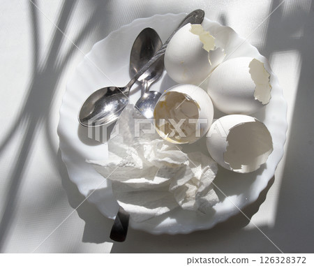 shells of boiled eggs with the remains of the yolk and the boiled egg white, used paper napkin, teaspoons, all on white plate, under the sun from window on a clean plastic windowsill with picturesque shells of boiled eggs with the remains of the yolk and the boiled egg white, used paper napkin, teaspoons, all on white plate, under the sun from window on a clean plastic windowsill with picturesque 126328372