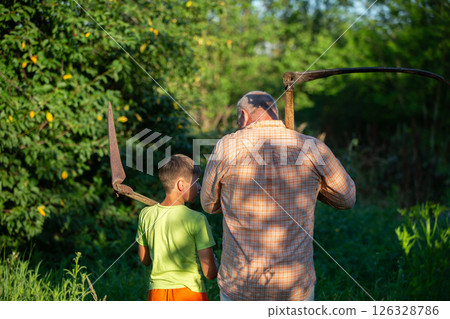 A man and a boy, father and son, work together in the field. The father with a scythe in his hands stands with his back to the camera. 126328786