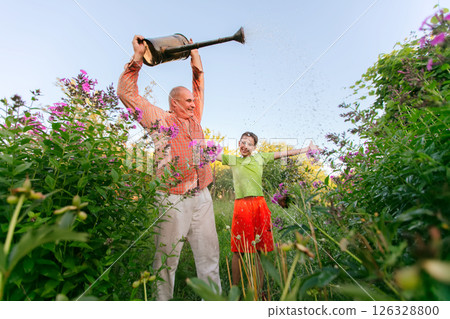 A man and a boy stand in a garden, the man holds a watering can and the boy laughs. The scene is carefree and joyful as the grandfather and grandson enjoy time together in the garden. 126328800