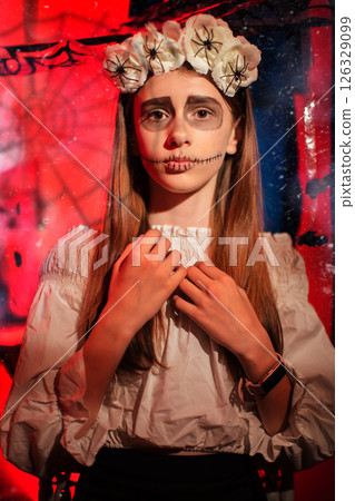 A teenage girl at a Halloween party with a skull painted on her face looks at the camera in fear. 126329099